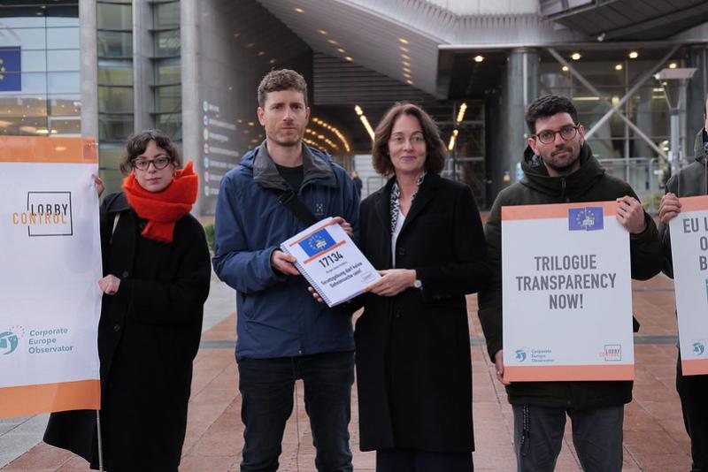 Four people stand in front of the European parliament. One holds a placard that reads "Trilogue Transparency Now"