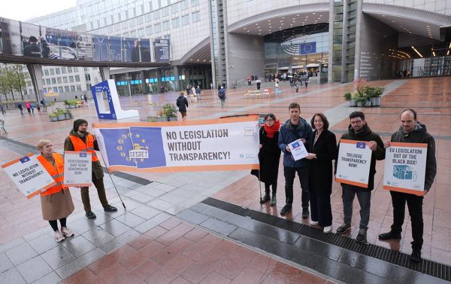Seven people stand in front of the European parliament holding placards and a banner that reads  "No legislation without transparency"